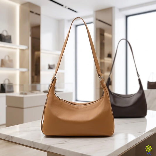 A tan leather shoulder bag with a single strap stands on a white marble counter in a modern boutique, with a darker brown version blurred in the background.