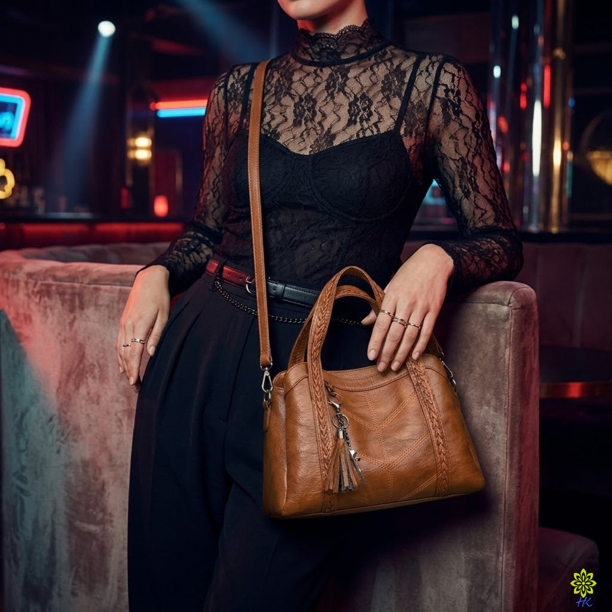 A woman wearing a black lace long-sleeve top and black pants carries a brown textured leather shoulder bag with a tassel charm while leaning on a velvet bench in a dimly lit, neon-lit environment.