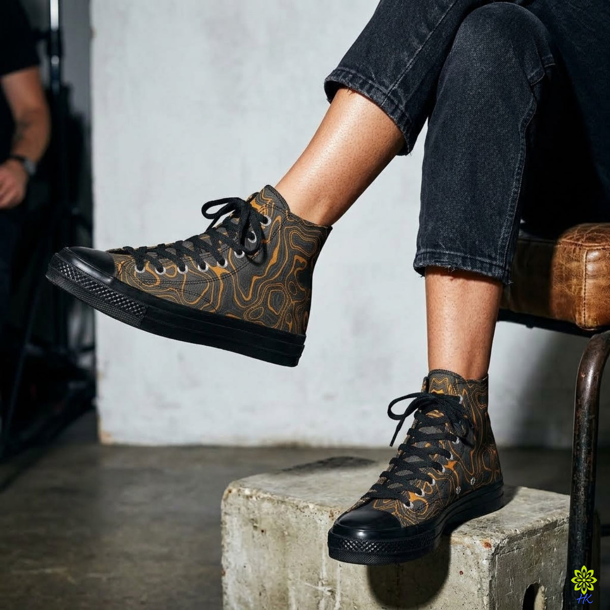 A person seated on a chair wearing brown high-top sneakers with a tan topographic map pattern and black soles, paired with dark cuffed jeans in an indoor setting.