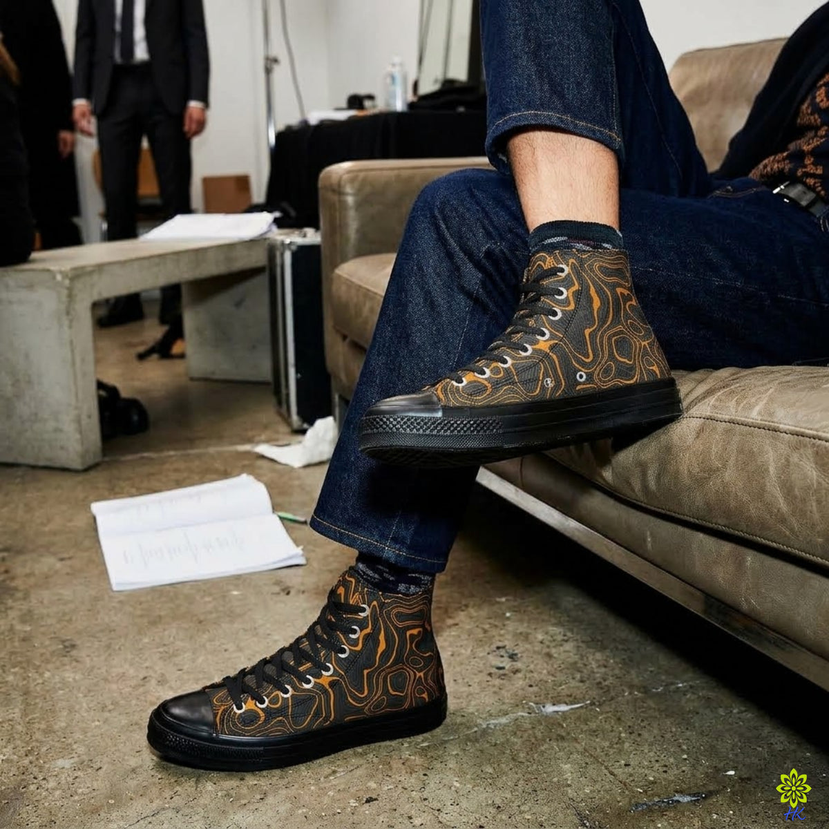 A person seated on a couch wearing black high-top sneakers with a brown and orange abstract flame-like pattern, paired with dark blue jeans in an indoor studio or backstage setting.