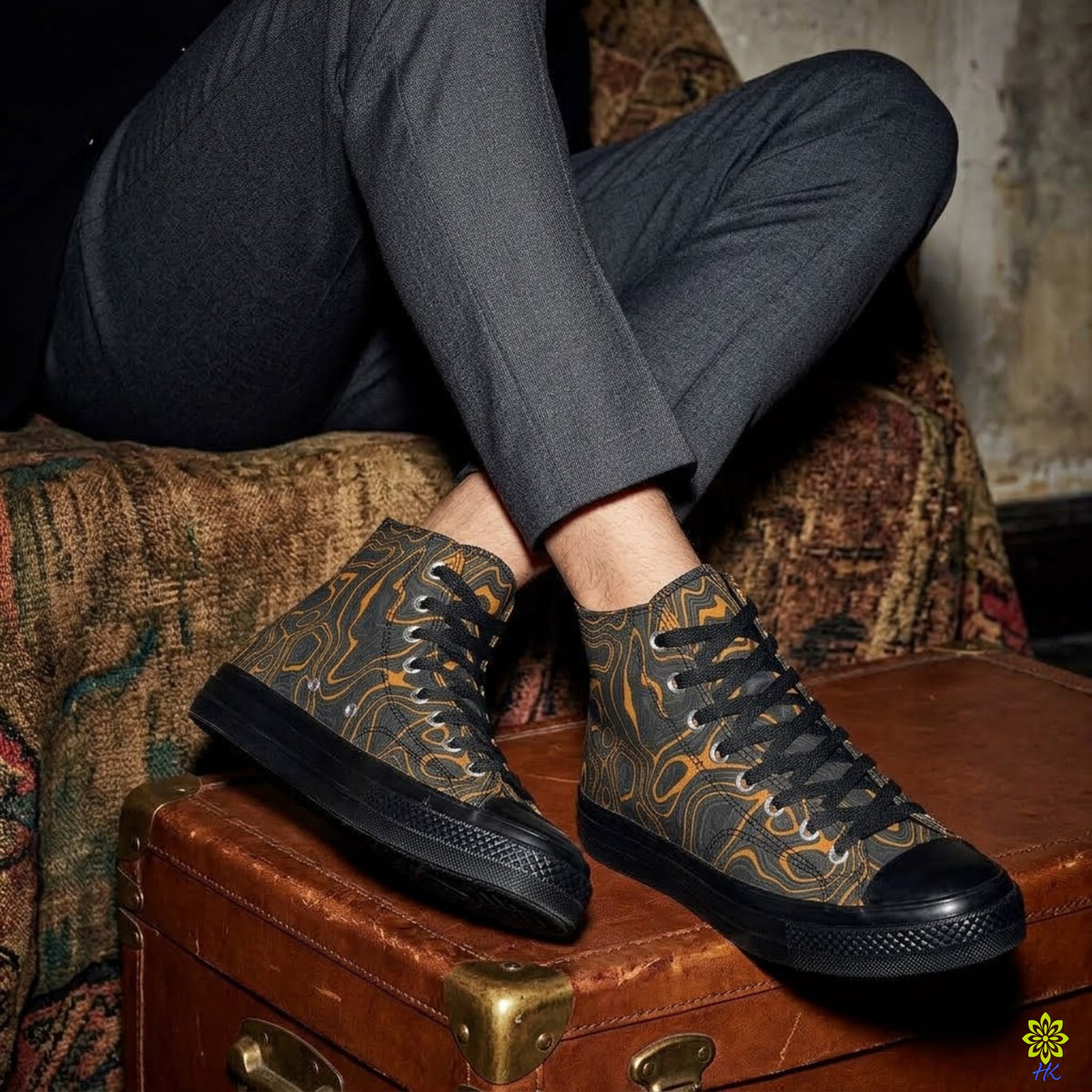 A person seated on a patterned armchair with crossed legs, wearing black high-top sneakers with a tan and brown abstract swirl pattern and black rubber toe caps. The shoes are shown resting on a vintage brown suitcase.