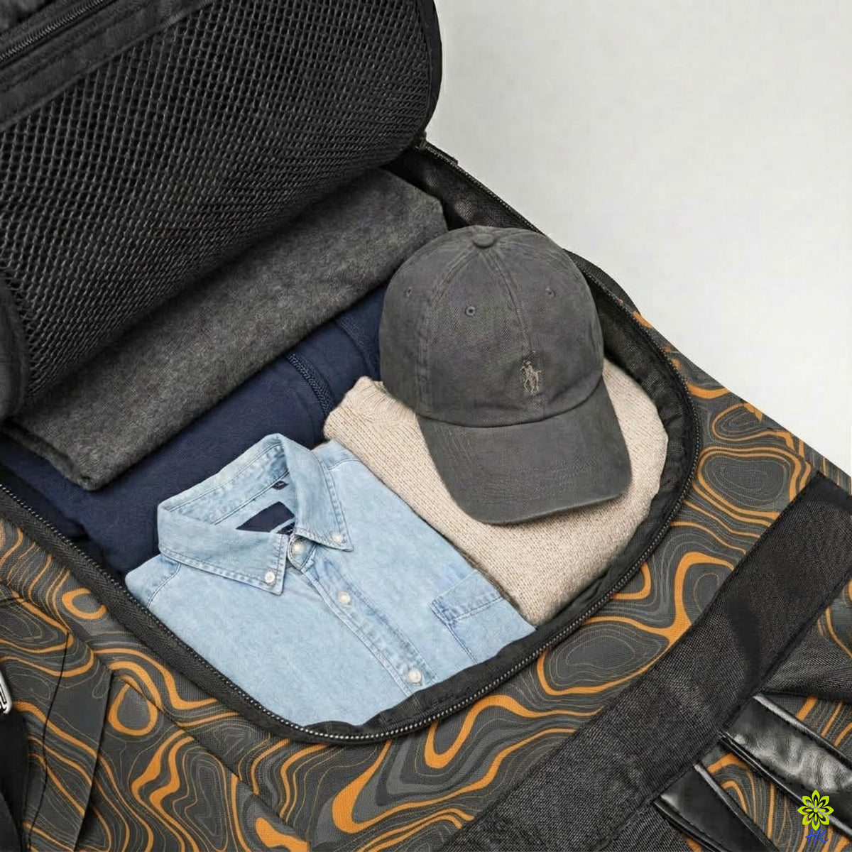 An open duffel bag with a black, gray, and orange wavy pattern, shown packed with folded clothing and a dark gray baseball cap.