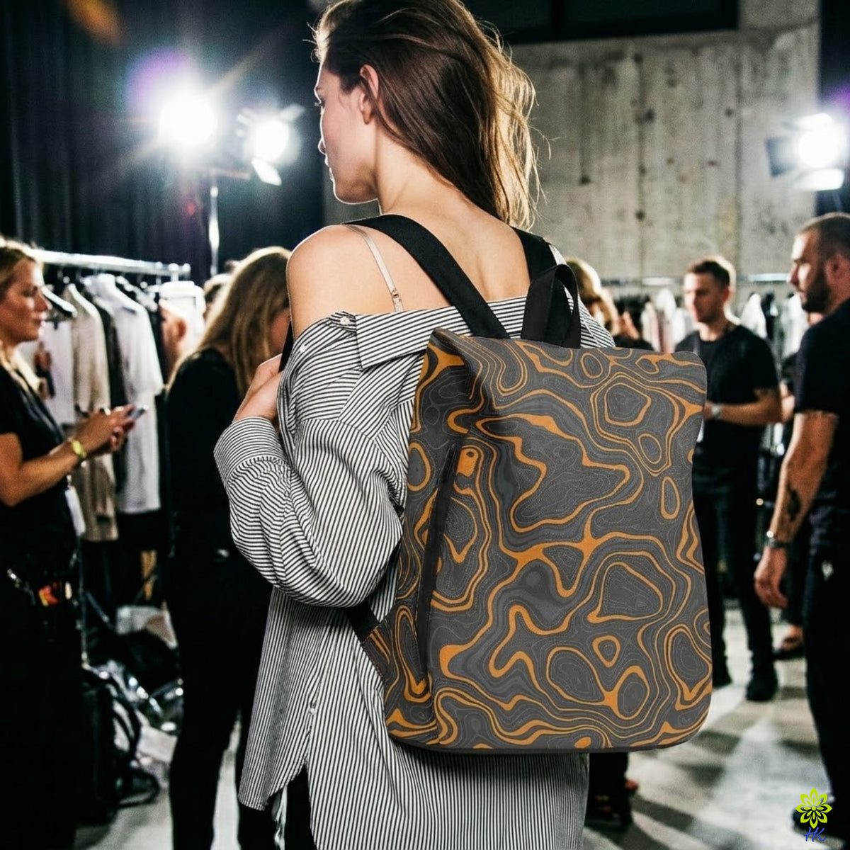 A woman in a gray and white striped shirt carries a large backpack with a gray and orange topographic contour pattern in a backstage fashion event setting.