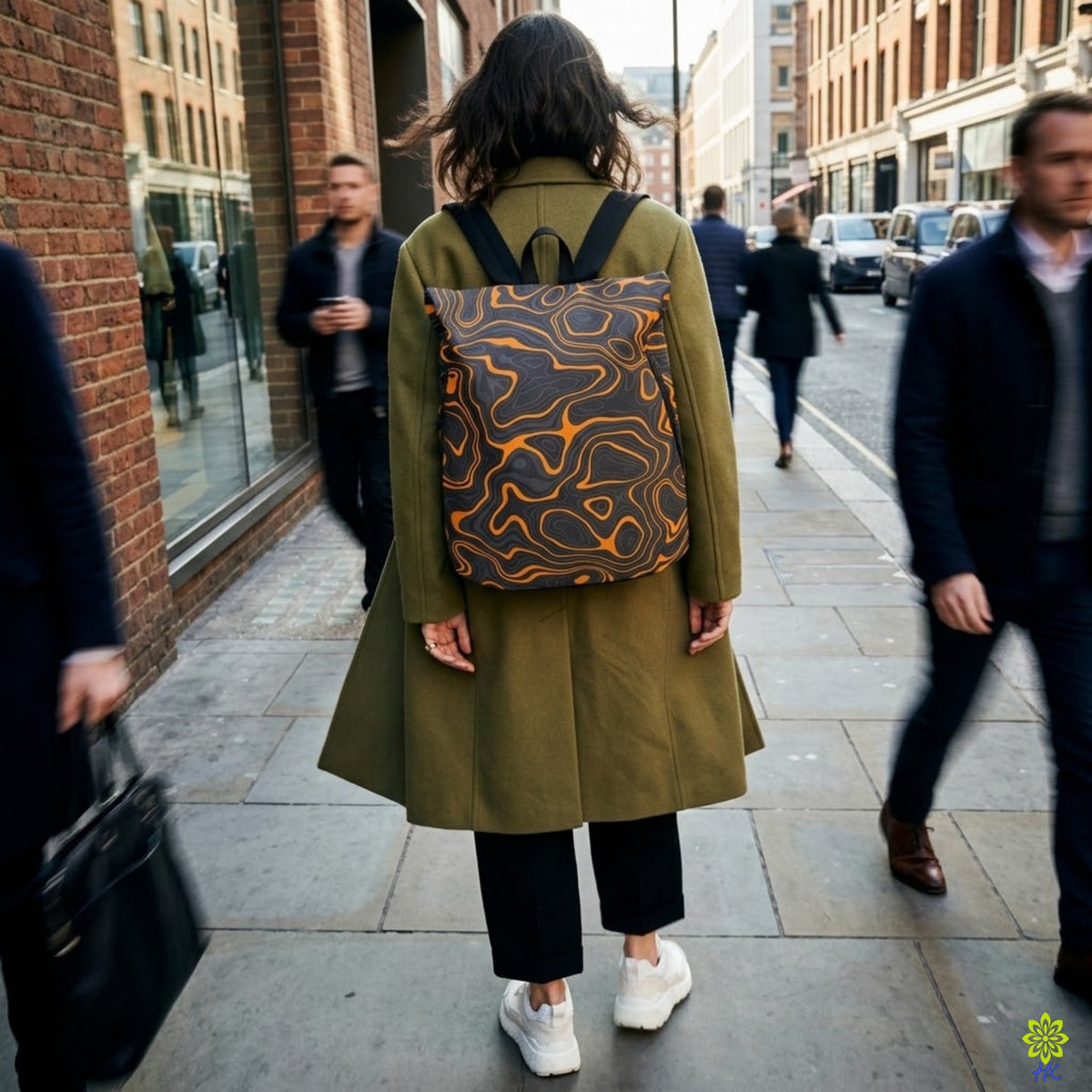 A woman seen from behind walking on a city sidewalk, wearing an olive green coat and a patterned backpack with orange and black wavy lines. Pedestrians and brick buildings are visible in the background.