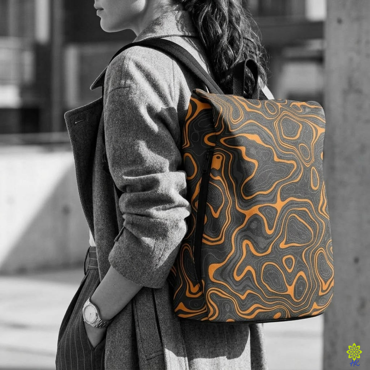 A woman in a gray coat carries a backpack with a black and orange topographic contour pattern, shown in a black and white lifestyle setting with selective color on the backpack.