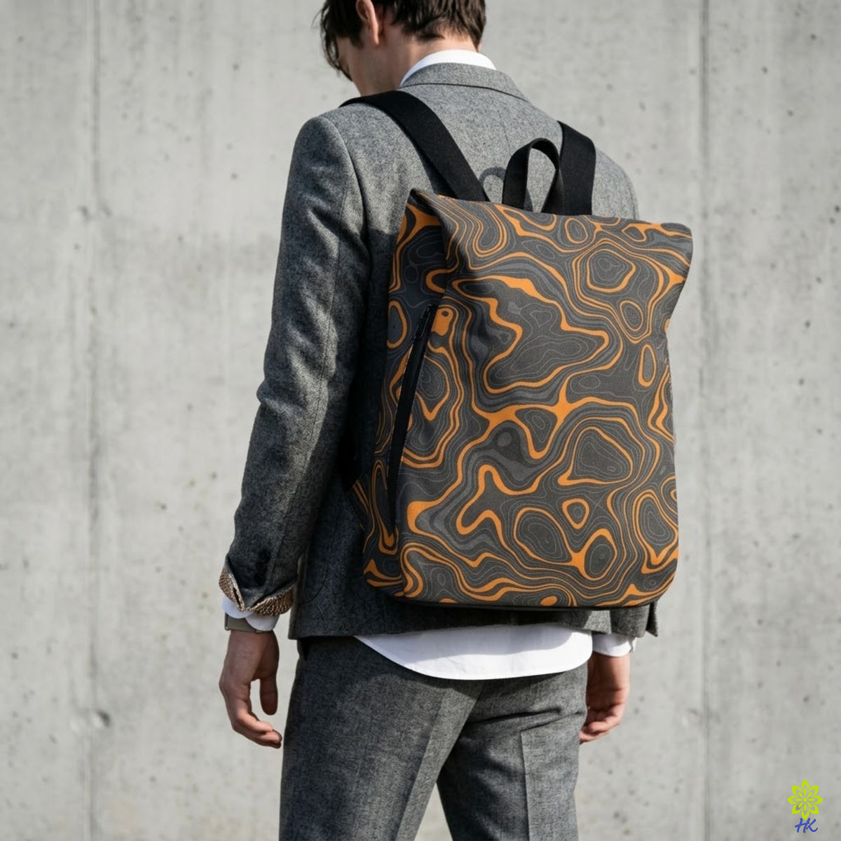 A man wearing a gray blazer and trousers is shown from behind, carrying a backpack with a black and orange topographic contour pattern against a concrete wall.
