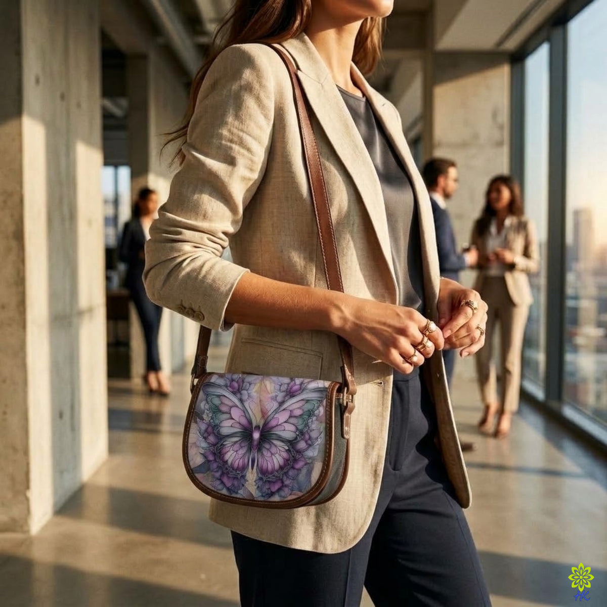 A woman in a beige blazer carries a small crossbody bag with a purple and blue butterfly print and brown leather trim, walking in a modern office hallway.