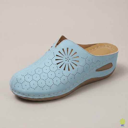 A light blue mule shoe with laser-cut geometric patterns on the toe and side, featuring a tan insole and sole edge, photographed on a neutral beige background.