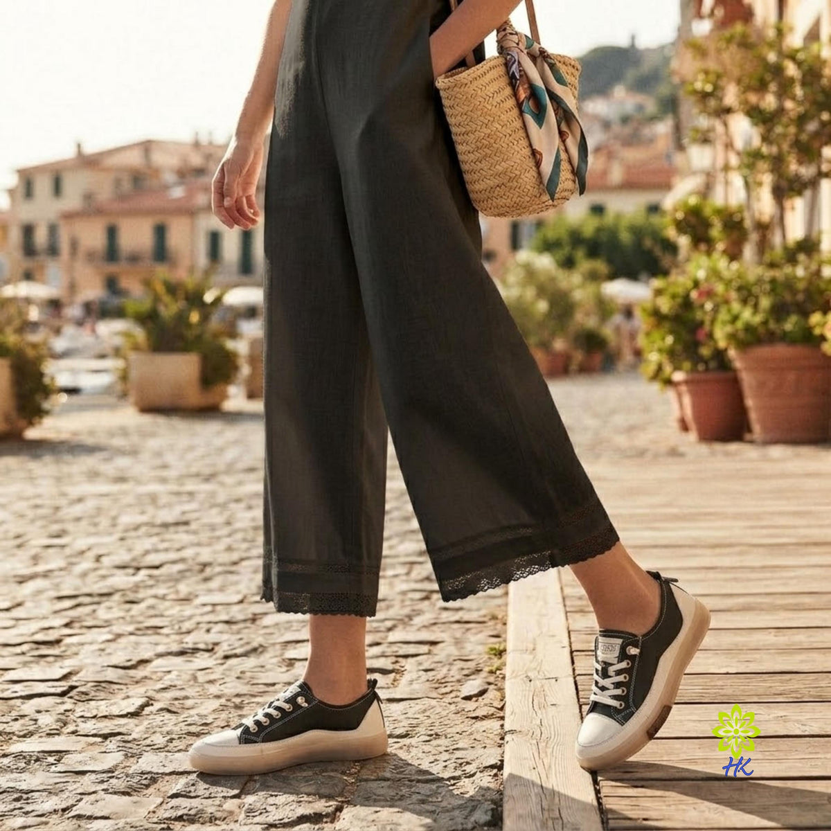 A person wearing dark charcoal wide-leg cropped pants with a lace trim at the hem, paired with black and cream low-top sneakers and carrying a woven straw tote bag with a patterned scarf, walking on a cobblestone street next to a wooden boardwalk.