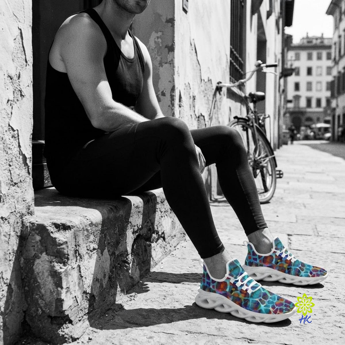 A man sits on a stone ledge in an outdoor street setting, wearing a black tank top and black leggings, with colorful patterned sneakers. A bicycle is parked in the background.