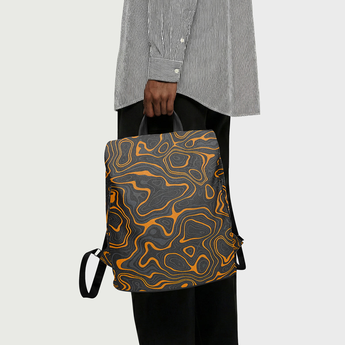 A person wearing a gray and white striped shirt and black pants holds a backpack with a dark gray and black topographic pattern outlined in bright orange, against a light gray background.
