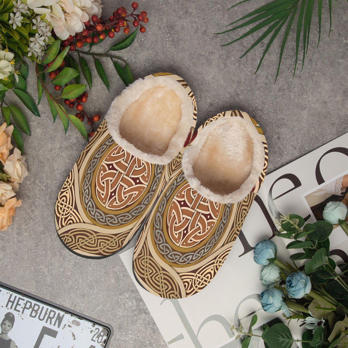 a pair of patterned slippers with fur lining, placed on a gray surface alongside various decorative elements such as flowers, berries, and a magazine.