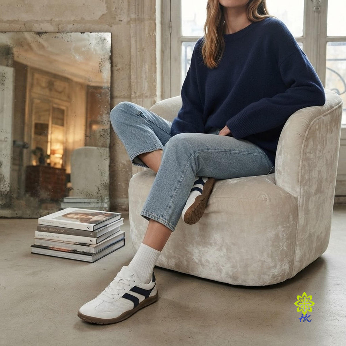 A woman sits in a cream-colored armchair wearing a navy blue crewneck sweater, light-wash cropped jeans, white socks, and white sneakers with navy stripes. The setting is a rustic room with a large mirror and a stack of books on the floor.
