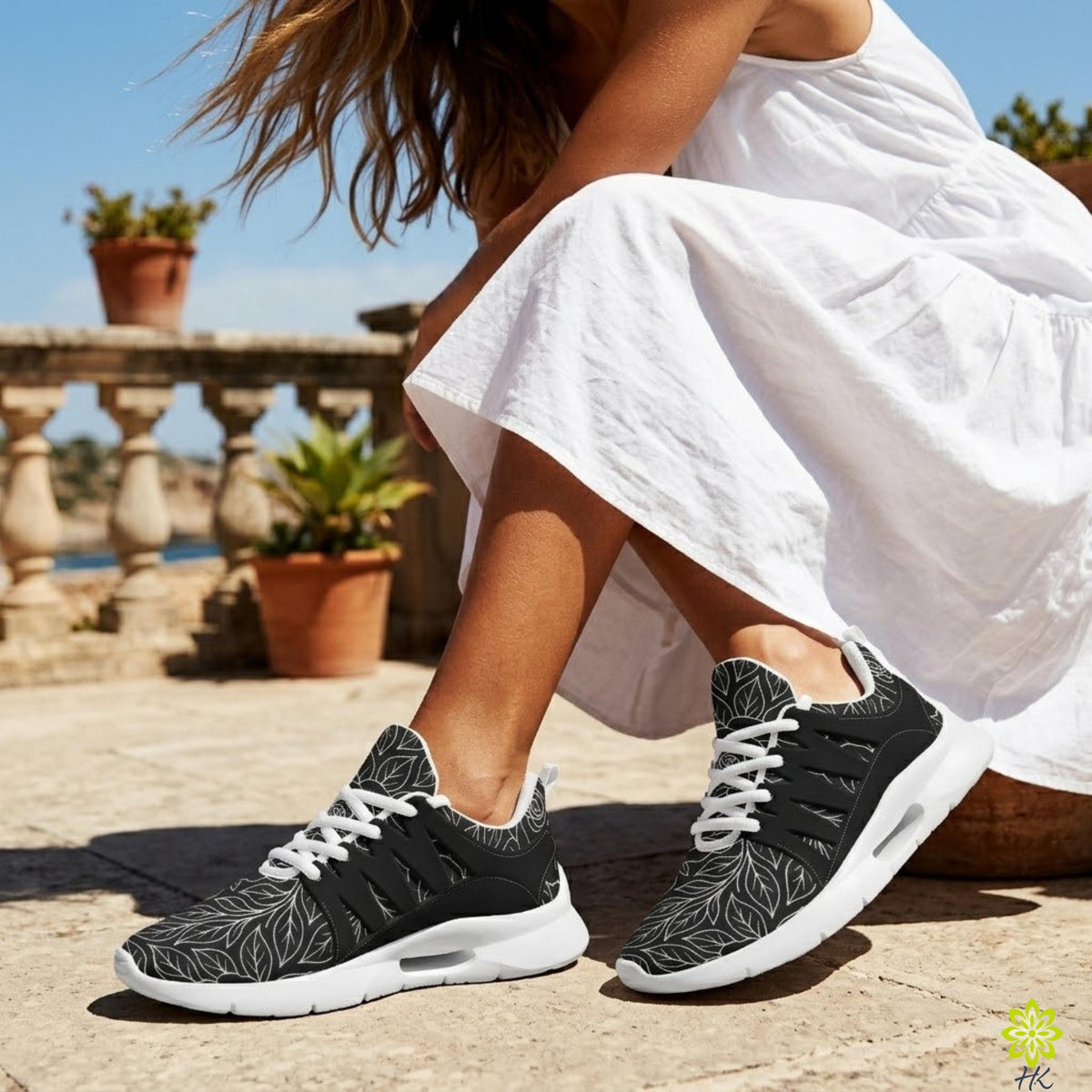 A woman wearing a white dress and black and white sneakers is sitting on a stone surface, with potted plants in the background.