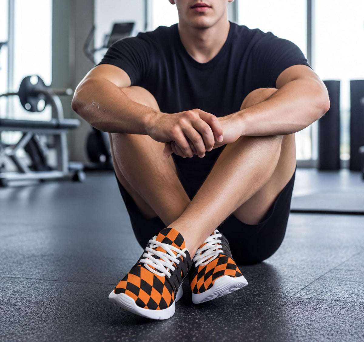 A man is sitting cross-legged on the floor in a gym, wearing black shorts and orange and black checkered sneakers.