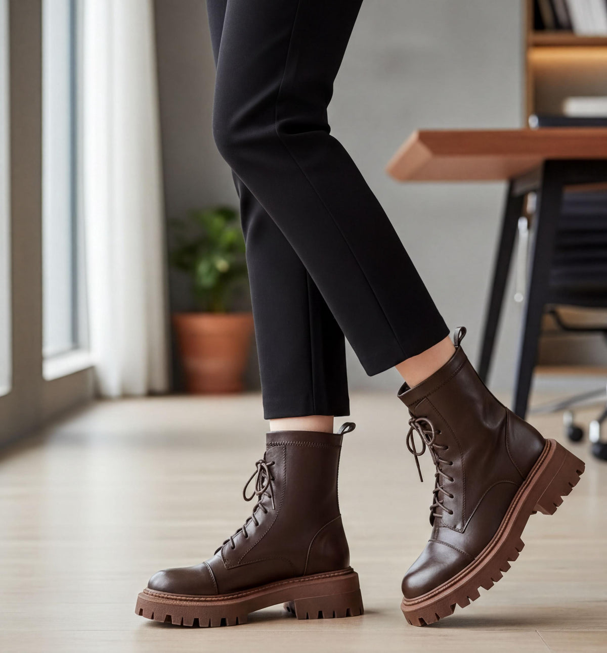 A person wearing black pants and brown boots stands in a room with a wooden desk and a window.
