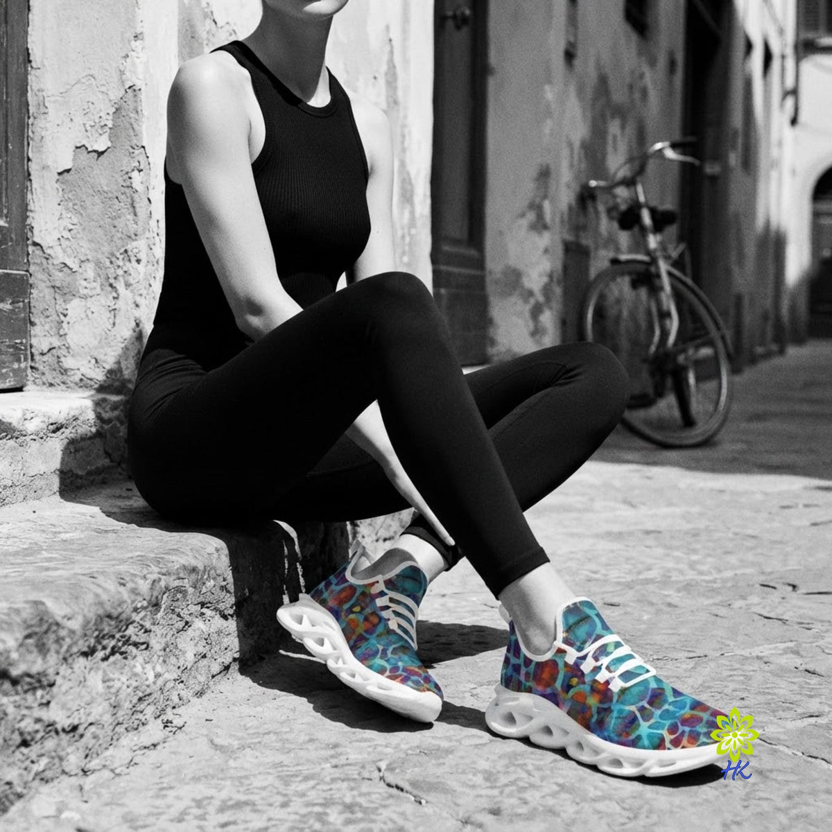 A person sitting outdoors on a stone ledge wearing a black sleeveless top and black leggings, with colorful patterned athletic sneakers. A bicycle and weathered building are visible in the background.