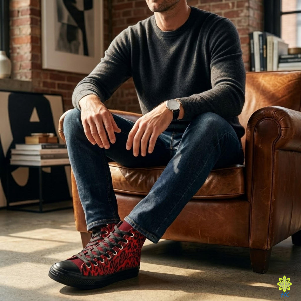 A man is sitting on a brown leather armchair, wearing a gray sweater, blue jeans, and red and black sneakers.