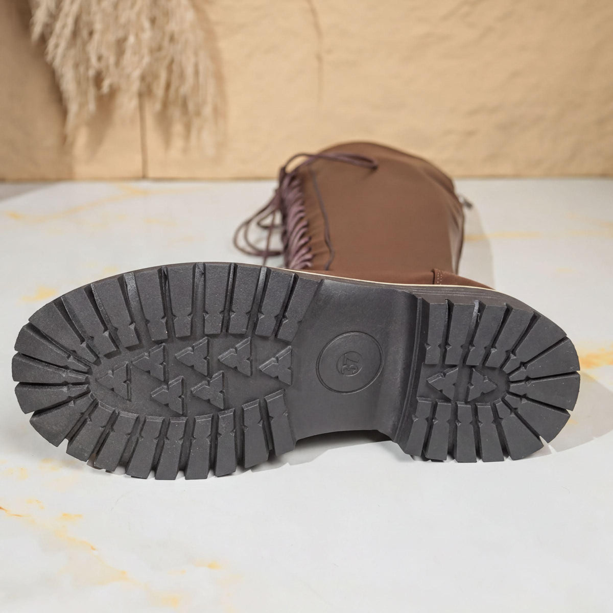 A close-up view of a brown leather boot with a distinctive tread pattern, placed on a white surface against a beige background.