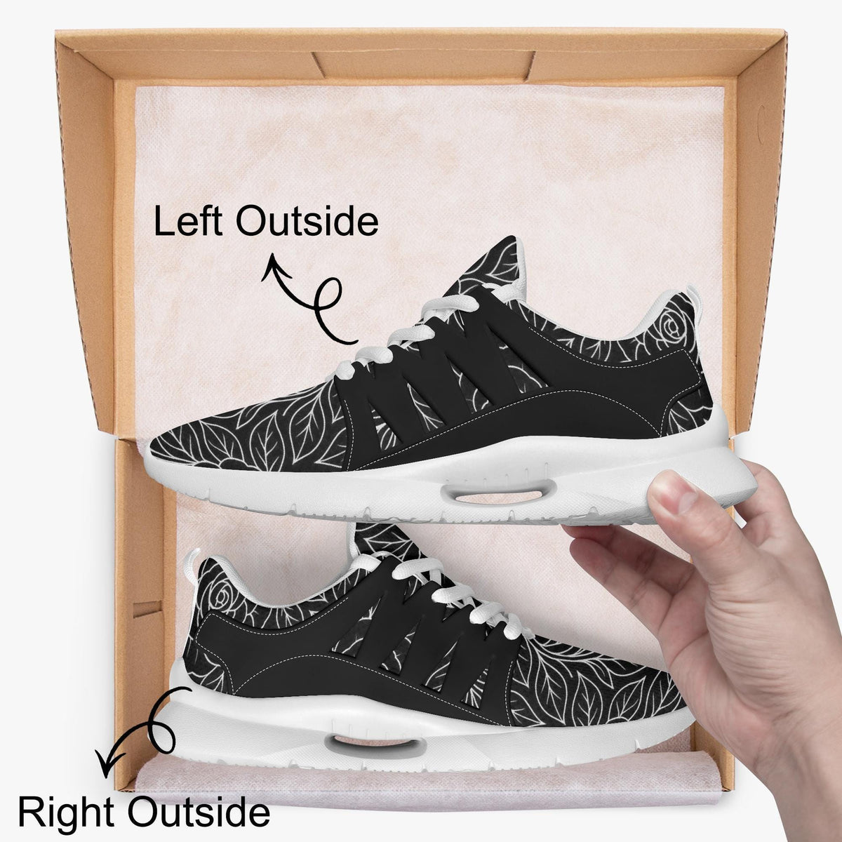 A hand is holding a pair of black and white sneakers with a floral pattern, standing inside a cardboard box.