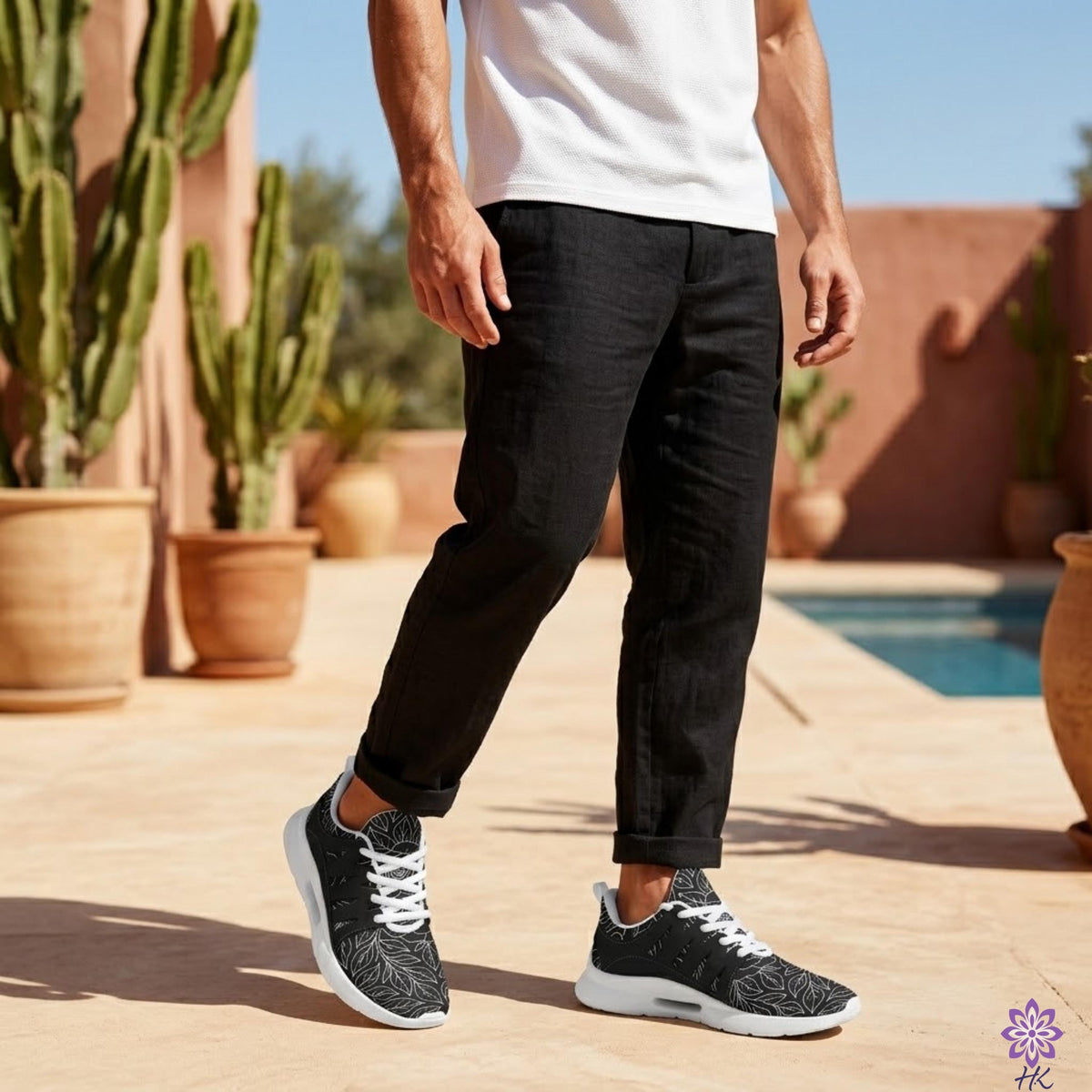 A man wearing black pants and black and white sneakers stands in front of a pool, with potted plants in the background.