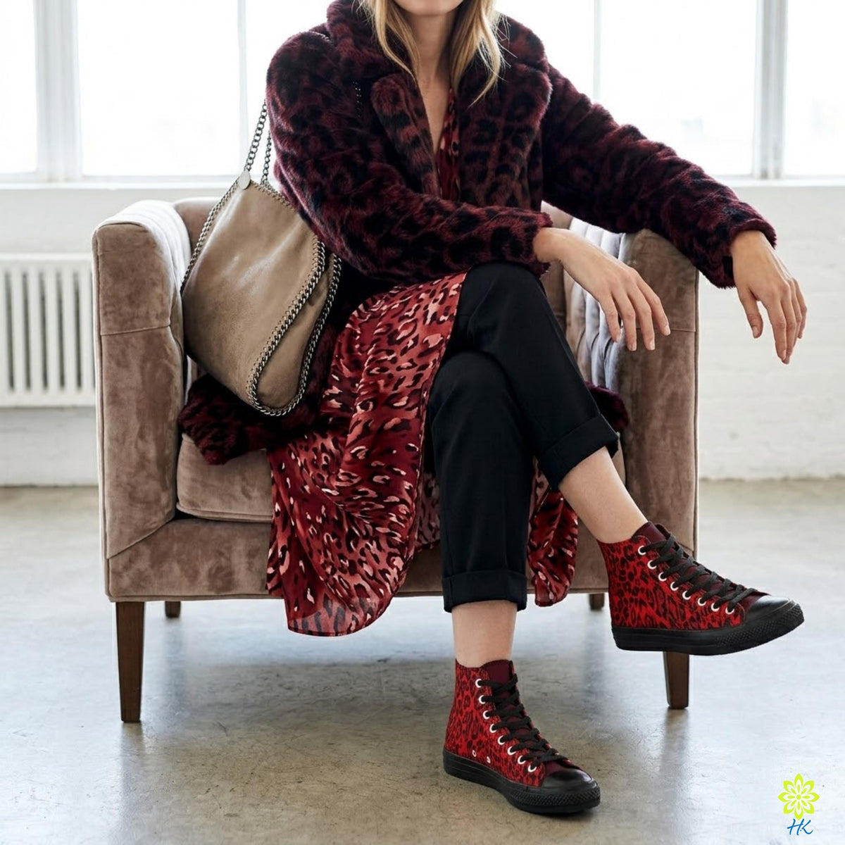 A woman wearing a burgundy coat, red and black leopard print dress, and red and black sneakers is sitting on a brown chair in a room with a window.