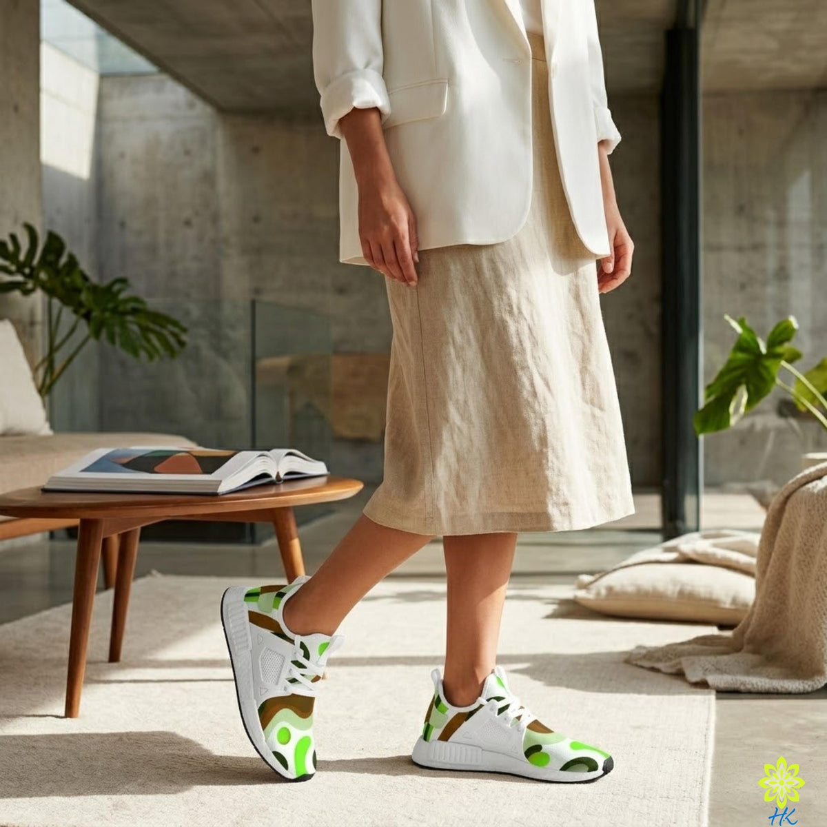 A person wearing a white blazer and beige skirt stands in a room with a wooden table, a plant, and a book.