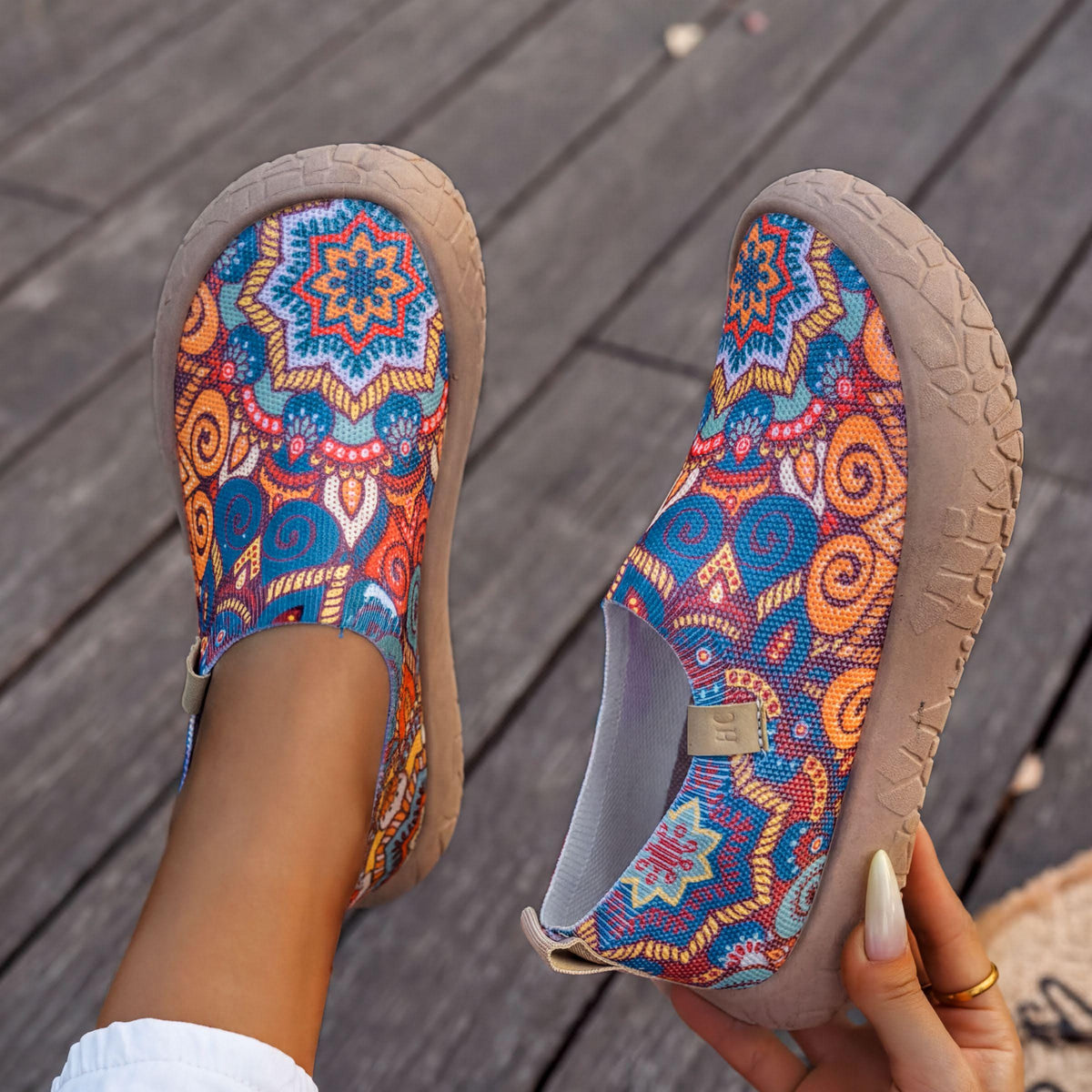 A pair of colorful, patterned slip-on shoes being held up by a person's hand.