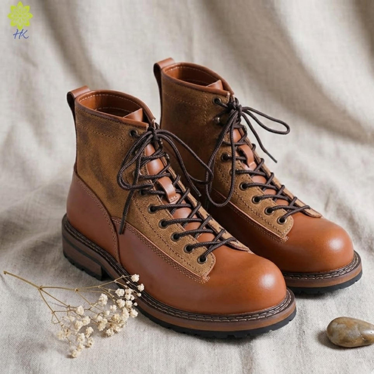 A pair of brown leather boots with laces and a thick sole, placed on a beige background with some white flowers and a small rock.