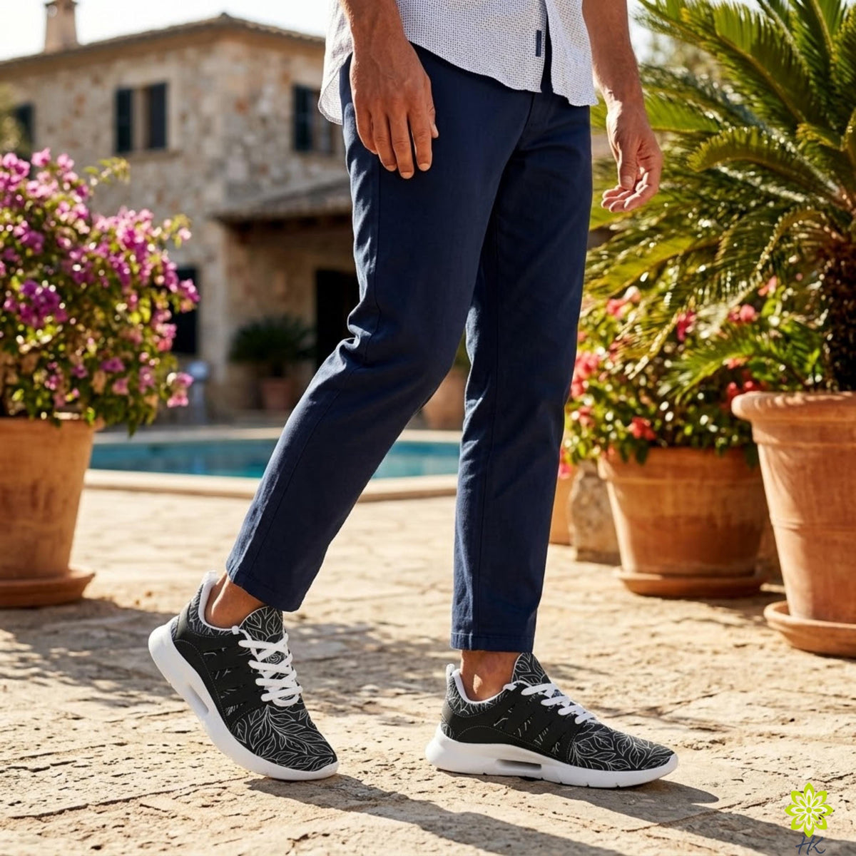 A person wearing navy blue pants and black and white sneakers stands in front of a house with a pool, surrounded by potted plants.