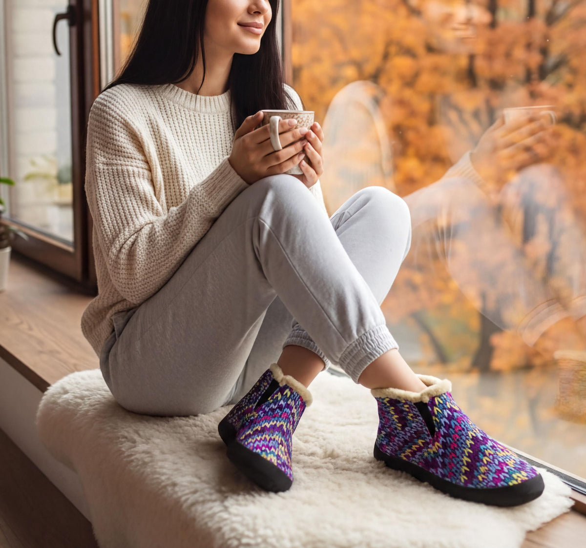 A woman sits on a window sill, holding a cup and wearing colorful patterned shoes, with a view of autumn trees outside.