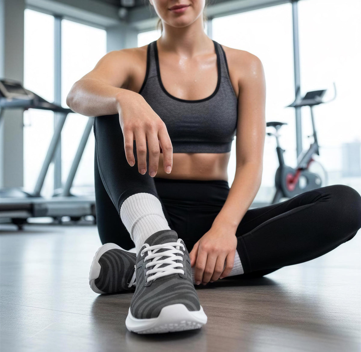 A woman in athletic clothing sits on the floor in a gym, with exercise equipment visible in the background.