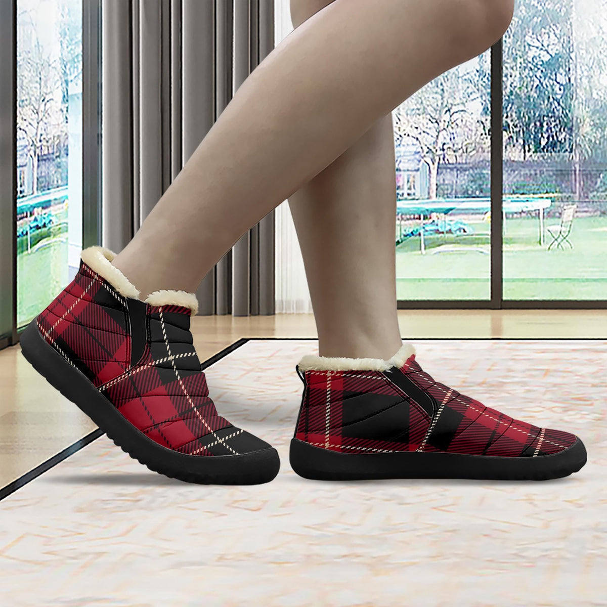 A person's feet wearing red and black plaid boots with fur lining, standing on a rug in front of a window with a view of trees outside.