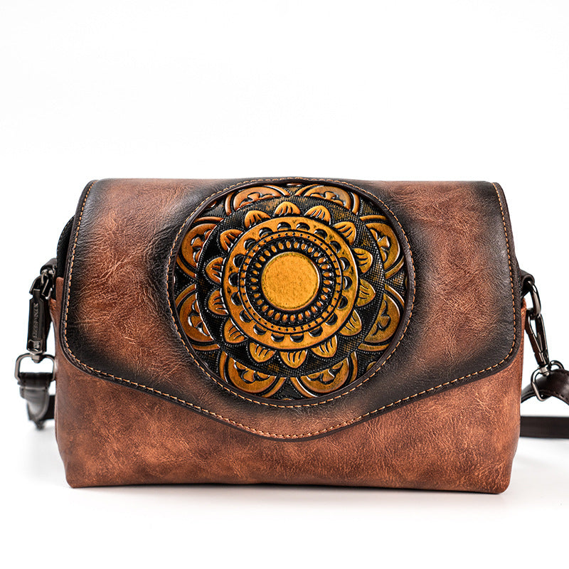 A brown distressed leather crossbody bag featuring a large circular floral mandala design in tan and black on the front flap, with a black shoulder strap visible on both sides.