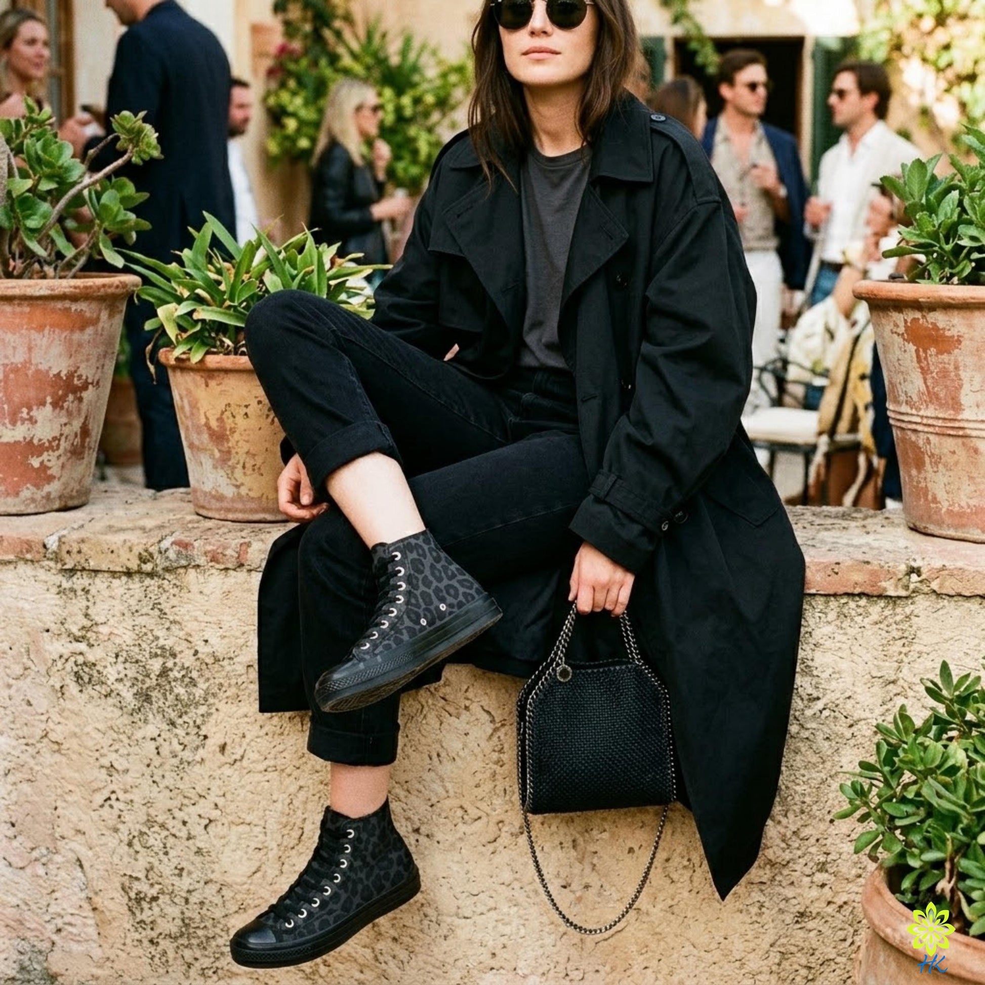 A woman in a black coat and sunglasses sits on a stone wall, holding a black handbag.