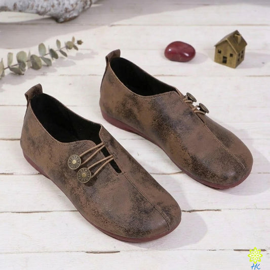 A pair of brown leather shoes with metal buttons and laces, placed on a white wooden surface alongside a small model house and some greenery.