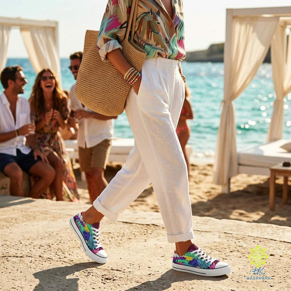 A woman wearing a colorful printed shirt and white pants walks on a beach, carrying a woven bag.