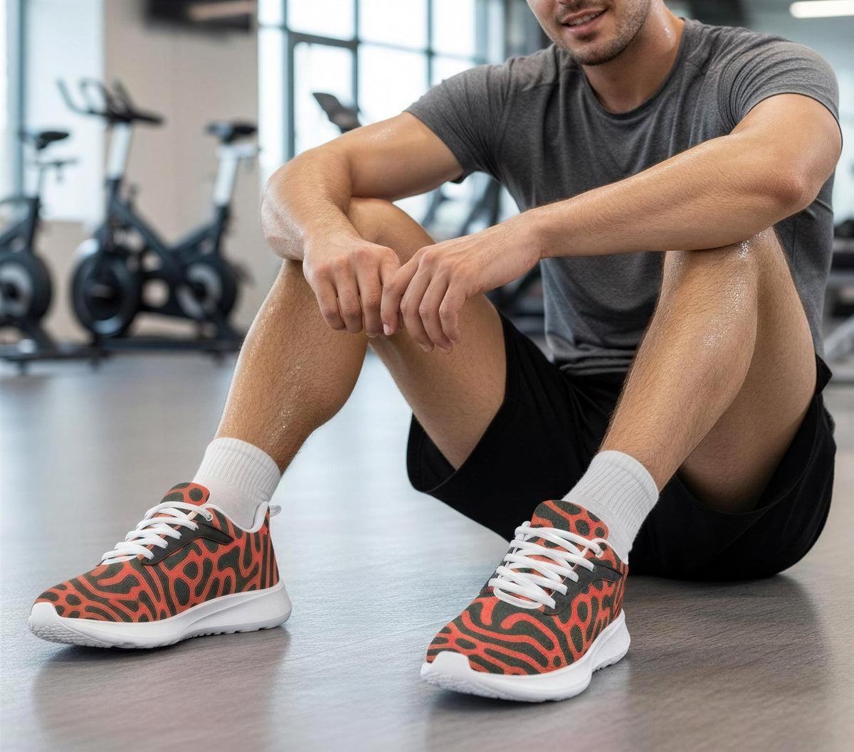 A man is sitting on the floor in a gym, wearing athletic shoes with a distinctive pattern.