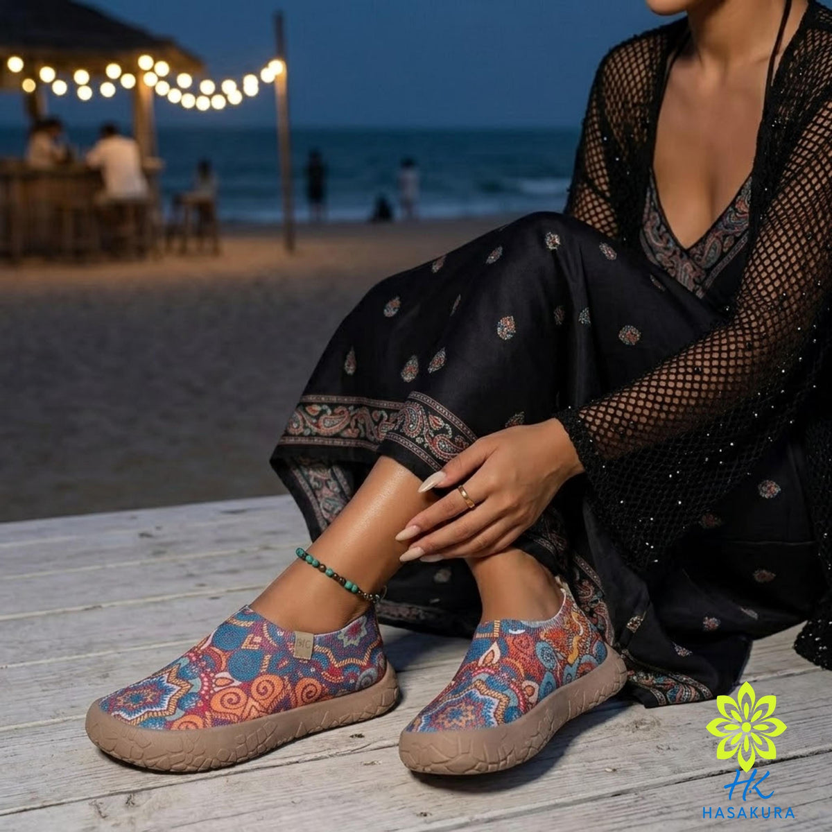 A woman in a black dress with a paisley pattern sits on a wooden boardwalk, wearing colorful patterned shoes and a black shawl. In the background, there is a beach with a string of lights and a wooden structure, creating a relaxing and festive atmosphere.