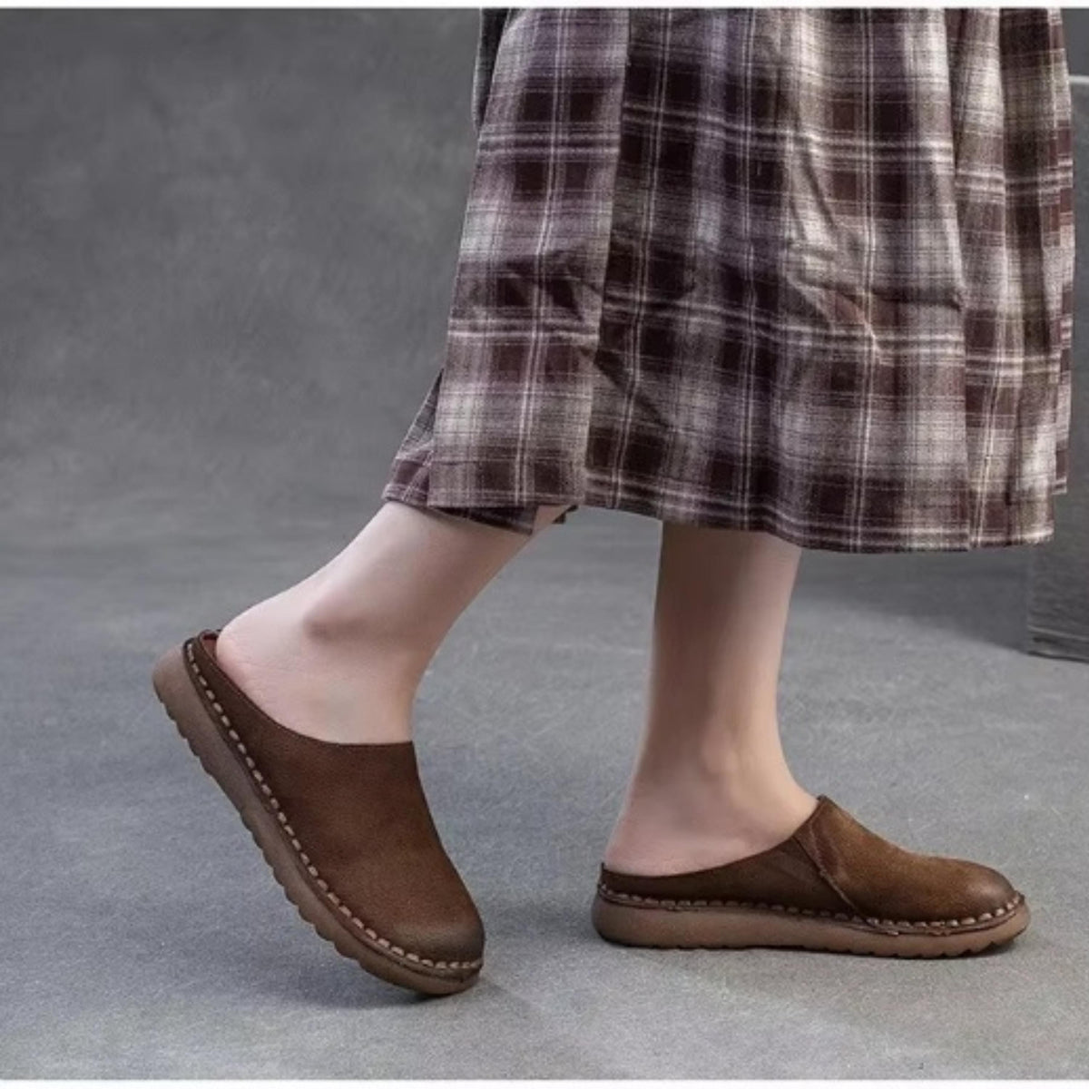 A person wearing brown leather shoes and a plaid skirt is standing on a gray surface.