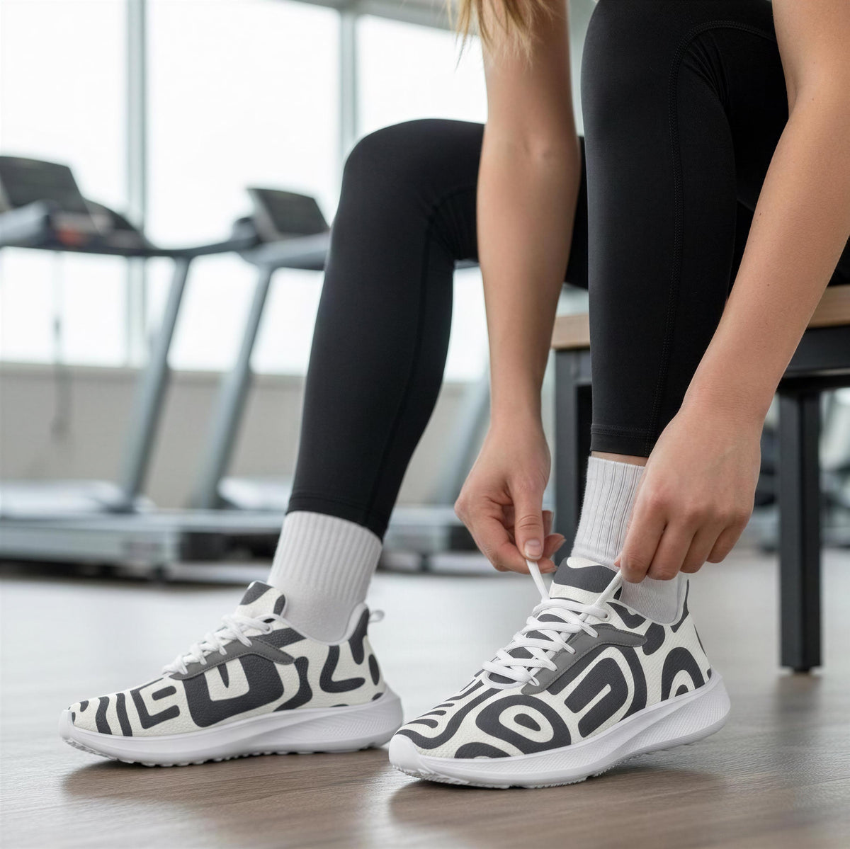 A person is tying their shoes in a gym, wearing black leggings and white sneakers with a black and white pattern.