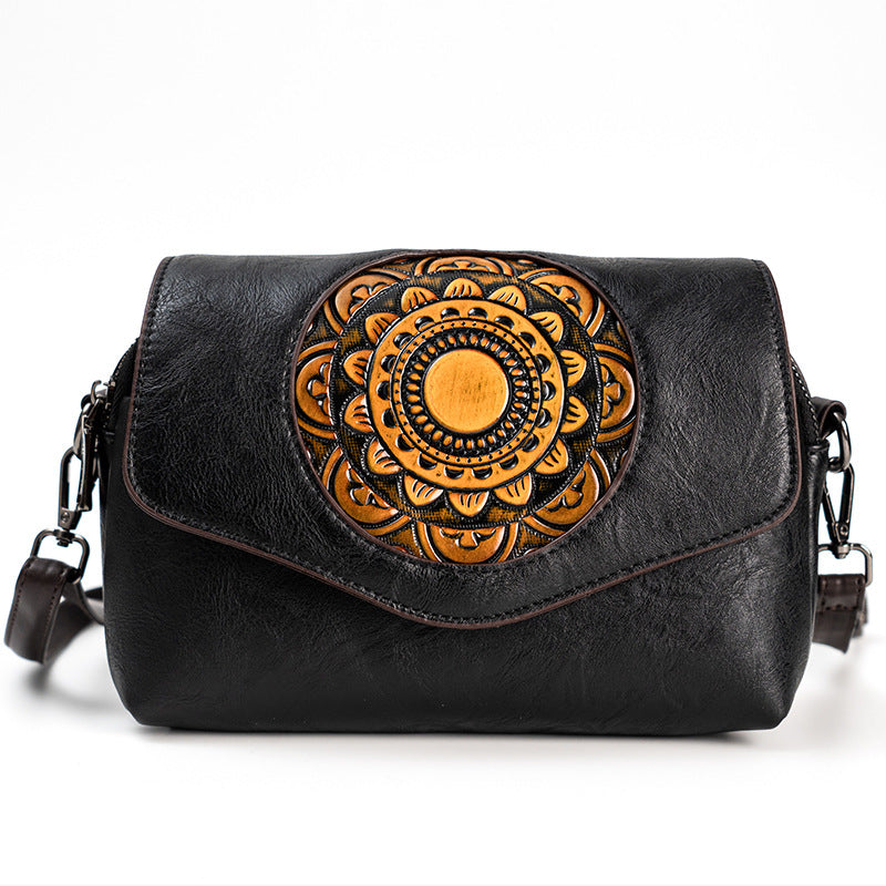 Black leather shoulder bag with a large circular tan and black tooled floral mandala design on the front flap, shown against a plain white background.