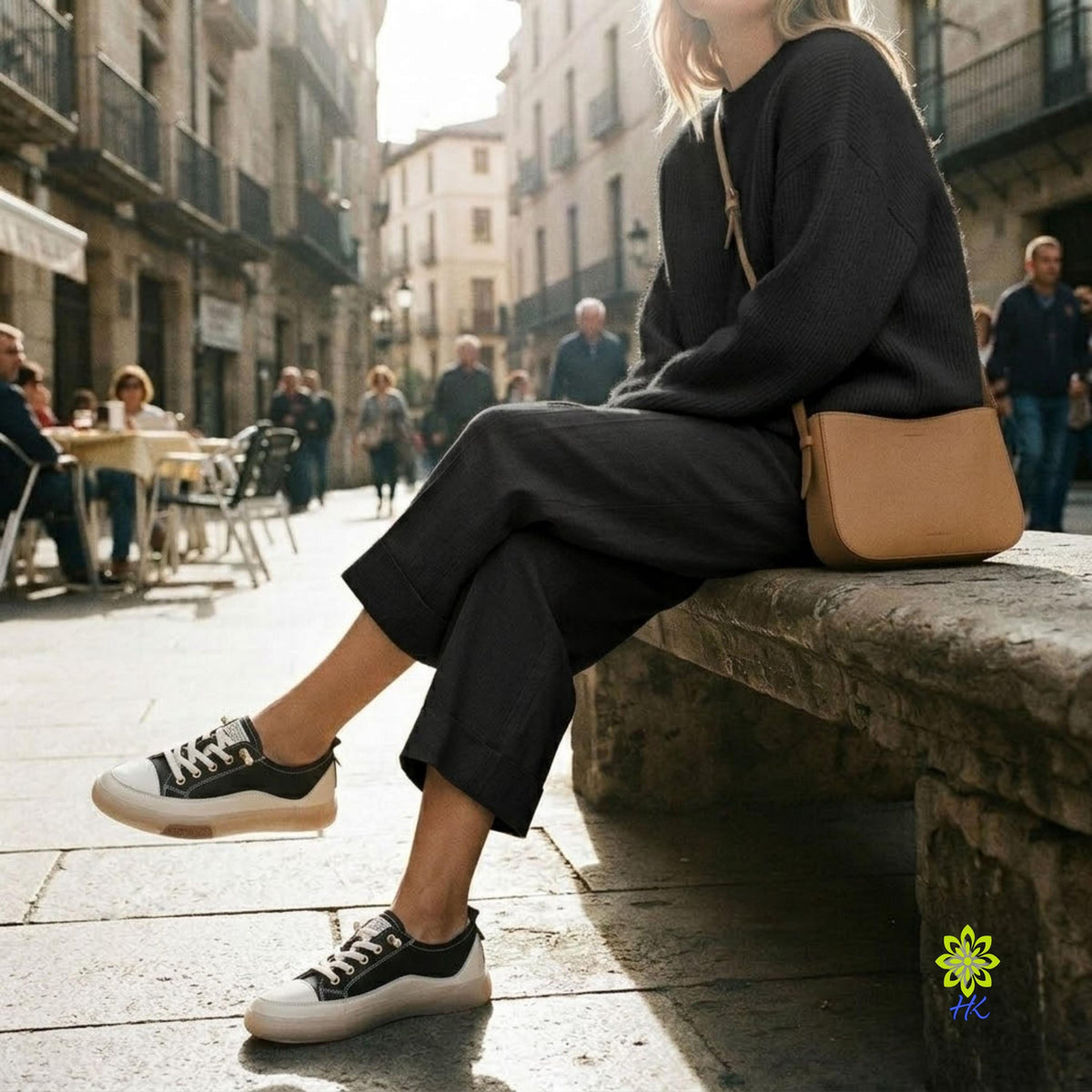 A woman sits on a stone bench in a sunlit city street, wearing a dark charcoal knit sweater and matching wide-leg pants with black-and-white low-top sneakers. A tan shoulder bag rests beside her.