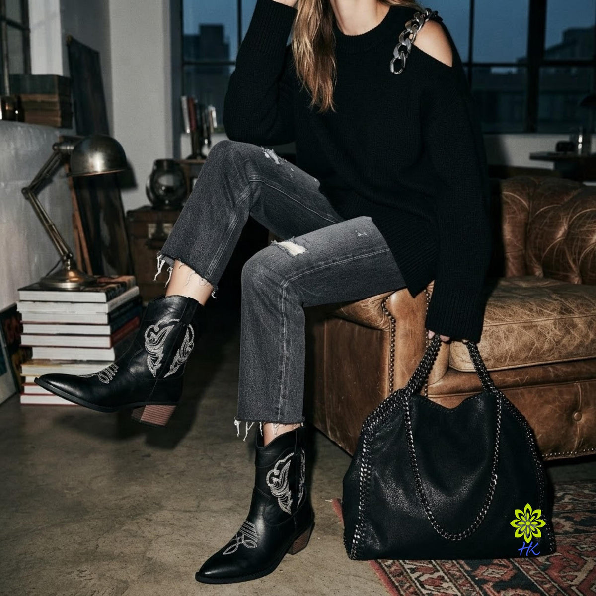 A person seated on a brown leather couch in a dimly lit room, wearing a black off-the-shoulder sweater, distressed gray jeans, and black western-style ankle boots with white embroidery. A large black leather handbag with chain straps rests beside them on the floor.